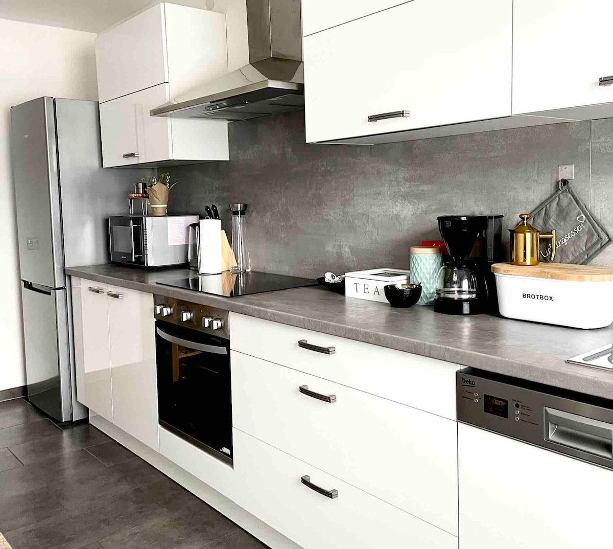 A modern kitchen is showcased, featuring sleek white cabinetry and stainless steel appliances. An organized countertop includes a coffee maker, kettle, and a decorative tea box. The refrigerator and built-in oven are visible, along with ample storage and workspace.