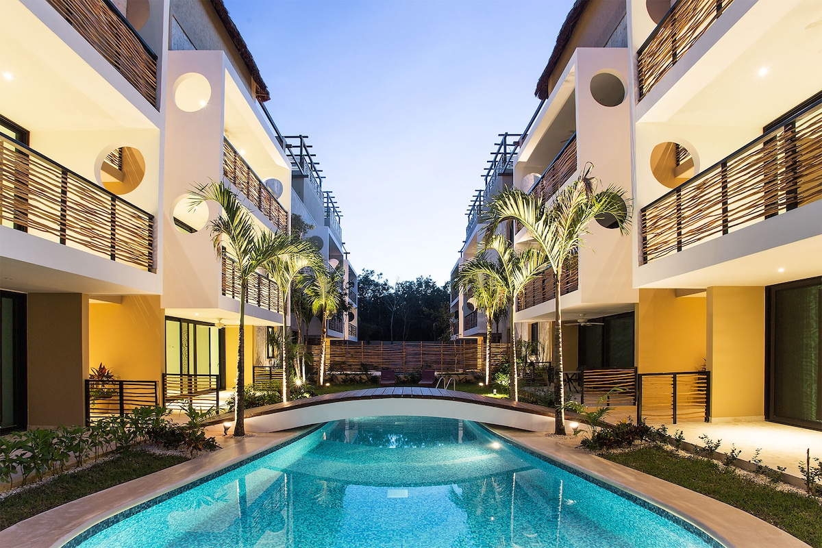 The image depicts a serene pool bordered by modern residential units. Lush greenery and palm trees frame the pool area, creating a calm atmosphere. Balconies with wooden railings provide a contemporary touch, while soft outdoor lighting enhances the inviting character of the space.