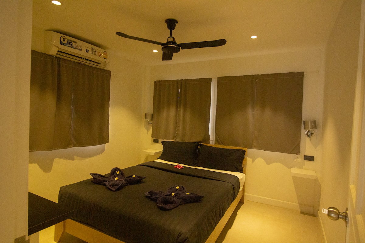 A comfortable sleeping area is featured with a spacious bed, accentuated by neatly arranged towels. Two windows with dark curtains allow for soft light, while a ceiling fan offers circulation. A minimalist and welcoming atmosphere is created with light-colored walls.