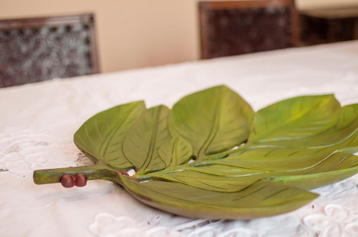 A decorative platter shaped like a large leaf is positioned on a white tablecloth. The platter features intricate leaf details in varying shades of green, with a stem extending on one side. The background includes blurred outlines of wooden chairs.