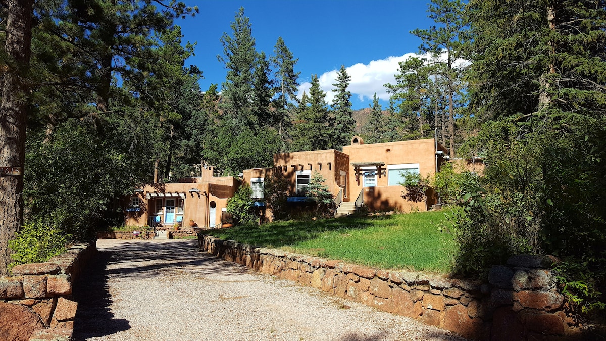 The exterior of the guesthouse showcases warm adobe-style architecture surrounded by pine trees and lush greenery. A gravel pathway leads to the entrance, and the distant mountains are visible under a clear blue sky.