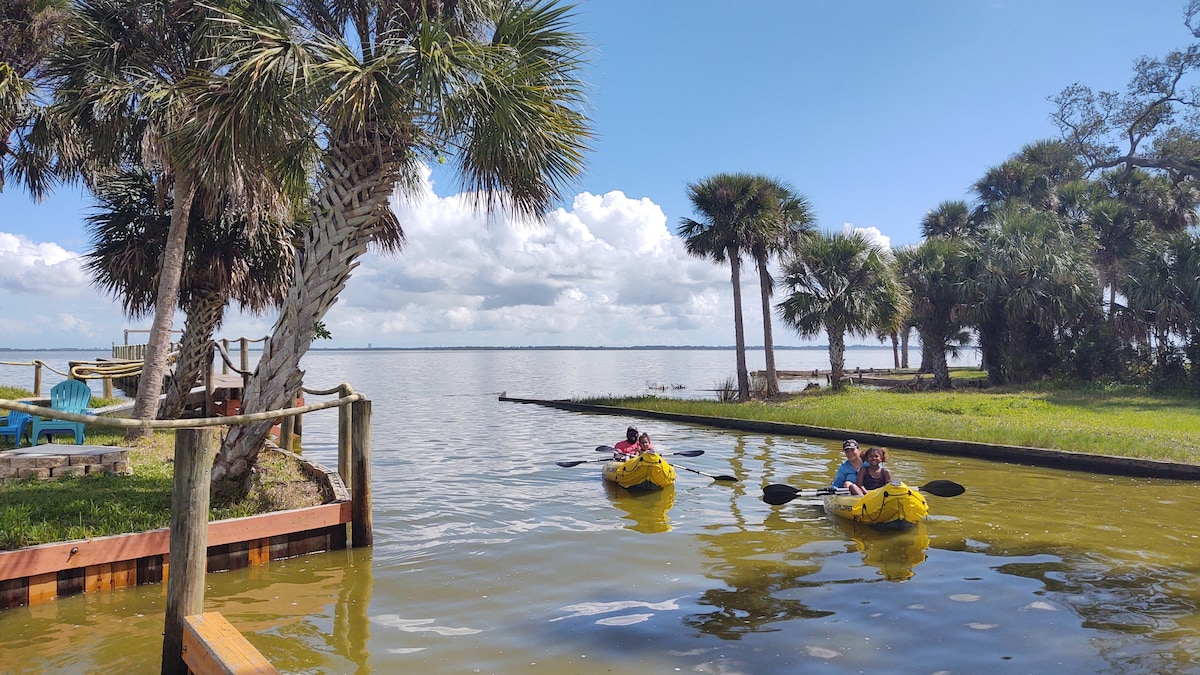 Two kayaks glide through calm water bordered by greenery and palm trees. The serene landscape features a wooden dock and blue skies with scattered clouds, setting a tranquil scene for kayaking on the waterfront.