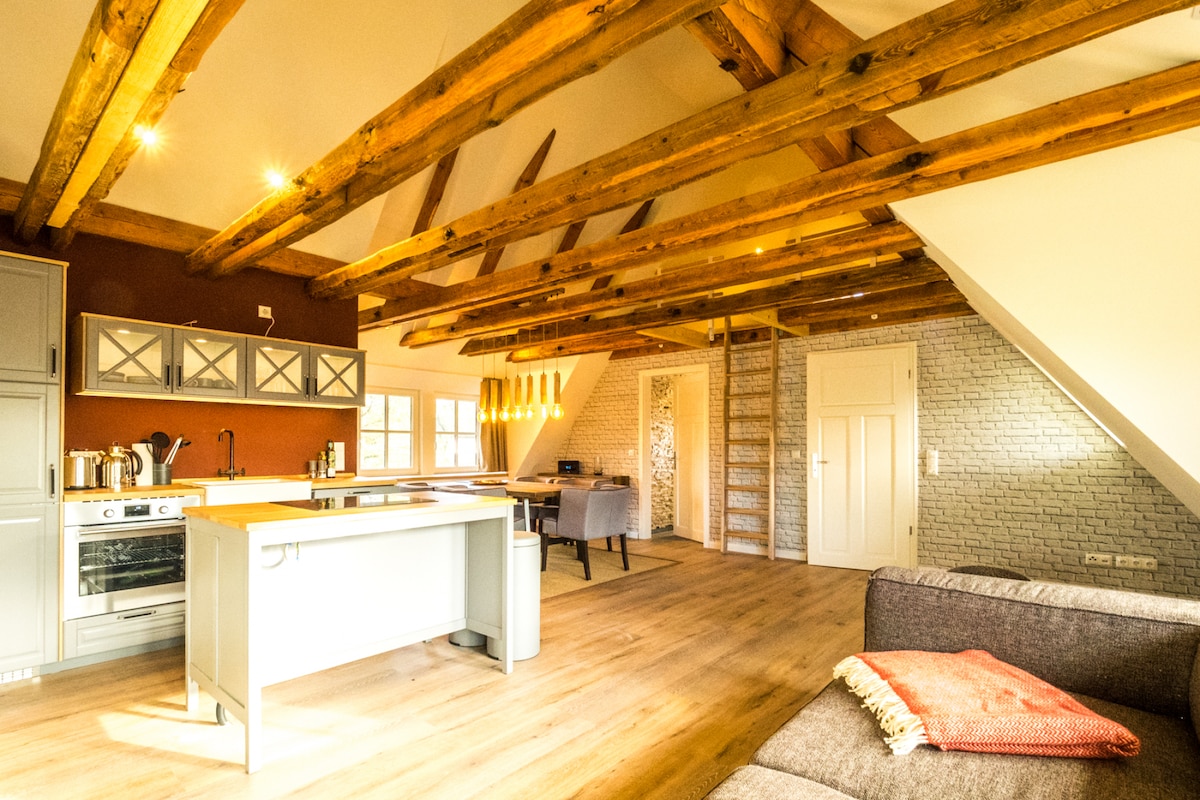 An open living space features exposed wooden beams and a mix of modern and rustic elements. A kitchen island with a stove is positioned centrally, surrounded by a living area with a cozy sofa. Light fills the room through large windows, enhancing the airy ambiance.