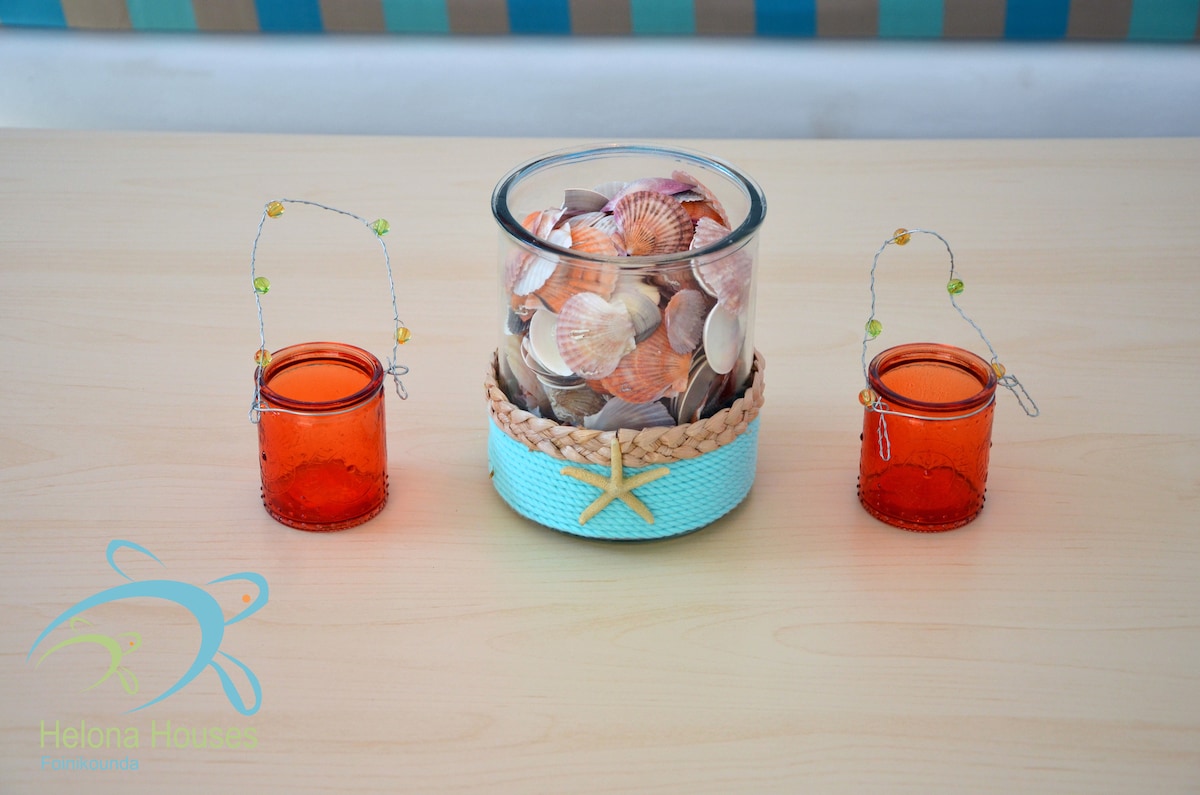 A decorative centerpiece is displayed on a light-colored table. It features a glass container filled with various seashells, bordered by a woven blue band. Two small red glass containers, each with wire handles, are positioned on either side, adding a charming coastal touch to the arrangement.