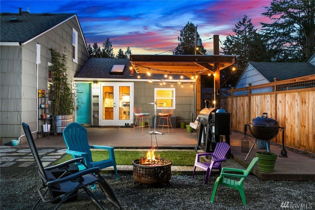 A private patio area features colorful seating options, including turquoise and purple chairs surrounding a fire pit. String lights hang above, illuminating the space. A barbecue grill and a dining area with chairs are visible, complementing the tranquil outdoor setting.