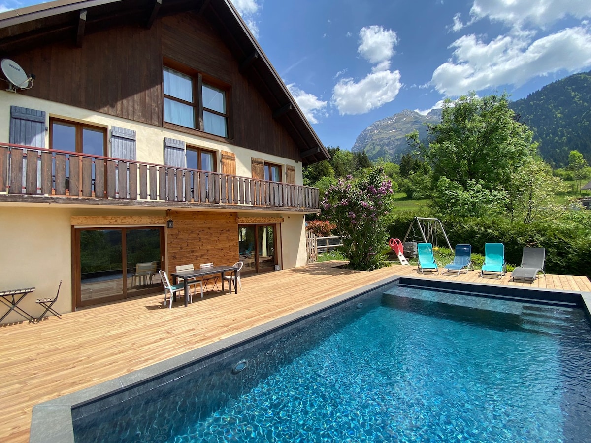 A spacious exterior view of a chalet features large windows and a wooden terrace. A crystal-clear pool is set amidst a well-maintained garden, with several lounge chairs positioned nearby. The backdrop includes lush greenery and majestic mountains under a bright blue sky.