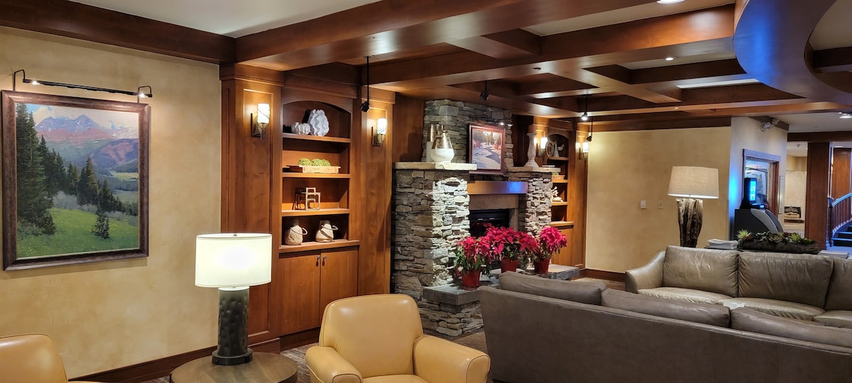 A welcoming lobby showcases warm wood paneling and natural stone accents around a central fireplace. Comfortable seating is arranged for relaxation, complemented by decorative shelves displaying art and local artifacts. Soft lighting enhances the cozy atmosphere, making it an inviting gathering space.