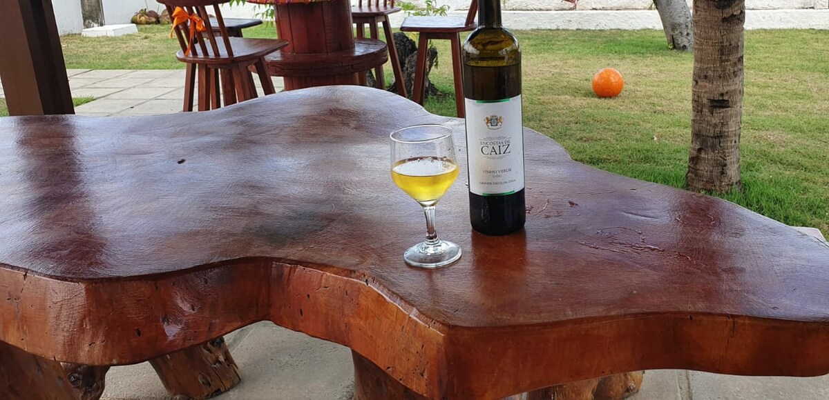 A natural wood table with a smooth, organic shape is displayed. A glass of light-colored beverage rests beside a bottle of wine. A grassy area and trees outline the scene, enhancing the outdoor setting.