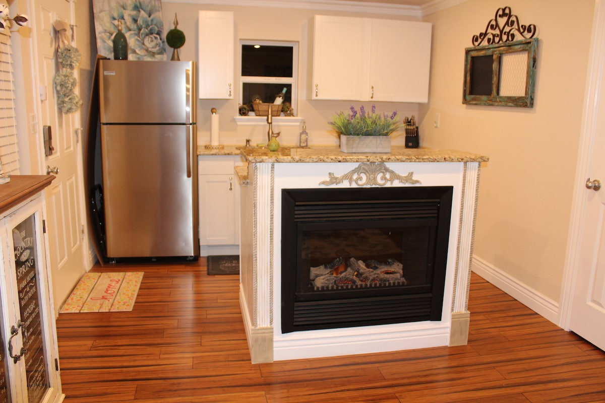 The kitchen showcases a stainless steel refrigerator next to white cabinetry and granite countertops. A decorative mantle surrounds the fireplace, which features a modern design. A window above the sink offers natural light, while plants add a touch of greenery to the space.