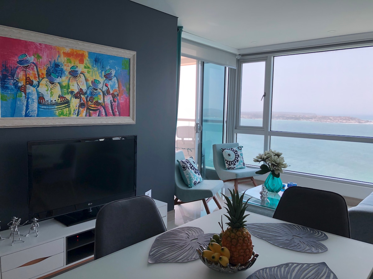 A modern living area features large windows with an ocean view, allowing natural light to fill the space. A colorful artwork is displayed on the wall, and a TV is positioned on a sleek console. A dining table with decorative fruit complements the stylish decor.