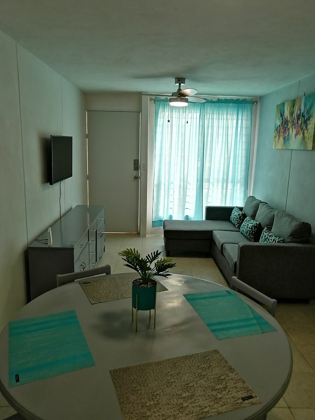 A light-filled living area features a gray sofa adorned with decorative cushions. A circular dining table is set with placemats, while a small potted plant adds a touch of greenery. Large windows draped with sheer curtains allow natural light to brighten the space.