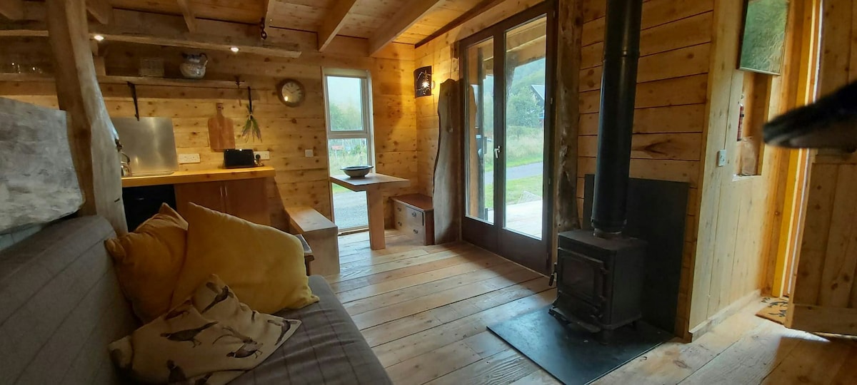 The interior of the cabin features warm wooden walls. A cozy sofa is adorned with colorful cushions. A wood burning stove sits on a stone base, providing a focal point. Large windows allow natural light, while a simple dining table awaits near the entrance.