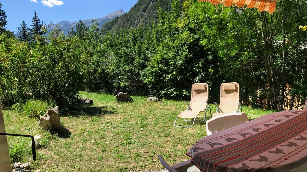 A spacious garden area is shown with a green lawn surrounded by trees and bushes. Two loungers are positioned to one side, inviting relaxation. A table with a striped cover sits in the foreground, offering a place for dining outdoors while enjoying the mountain views.