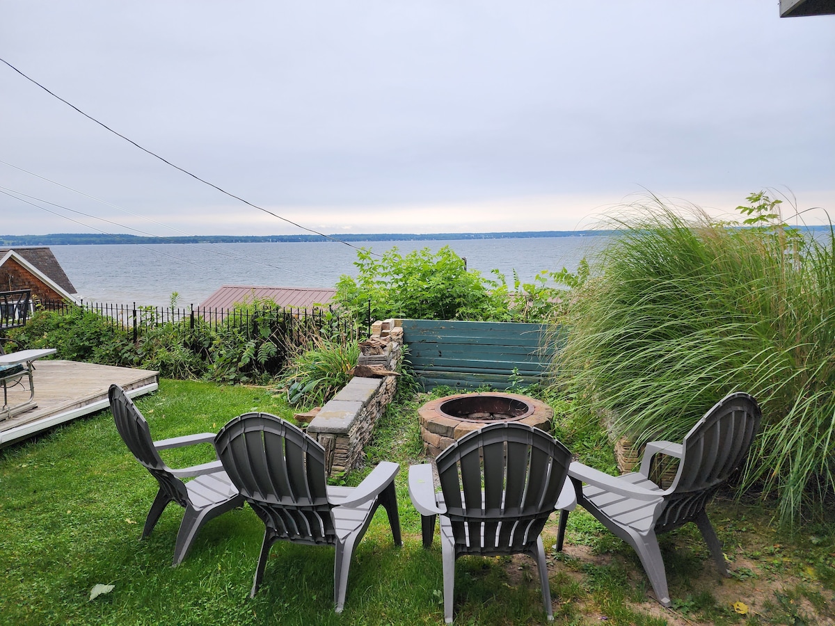 A serene outdoor space features four gray lounge chairs arranged around a circular stone fire pit. Overlooking the water, lush greenery and a wooden fence create a natural barrier, enhancing the peaceful setting. The cloudy sky reflects softly on the lake's surface.