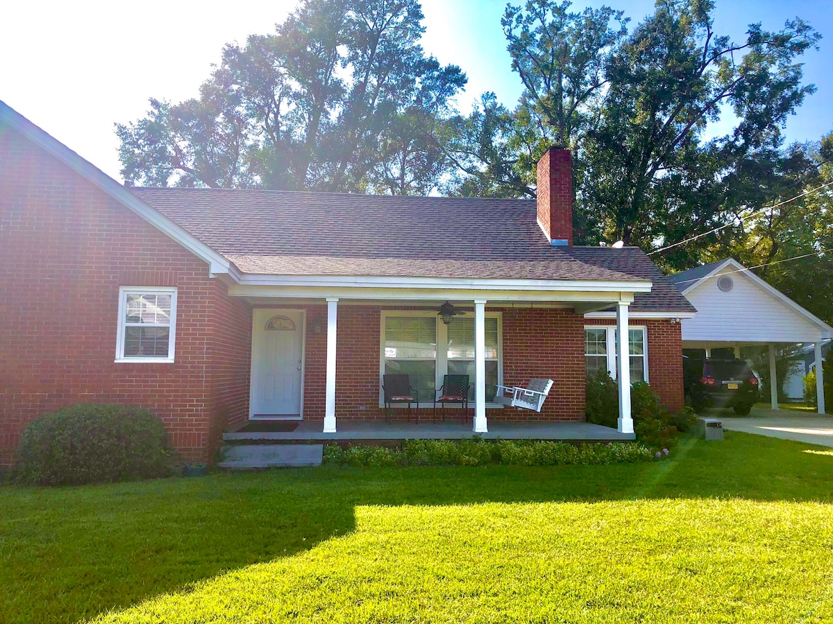 A charming brick house features a welcoming porch with comfortable seating and a classic rocking chair. Lush green grass surrounds the home, and tall trees provide natural shade in the background.