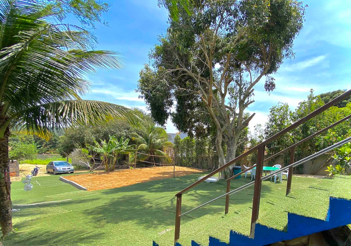 A spacious outdoor area is showcased, featuring well-maintained green grass and several tall trees providing shade. A volleyball court is visible along with a small seating area, complemented by a vehicle parked nearby. Blue stairs lead into this inviting space.