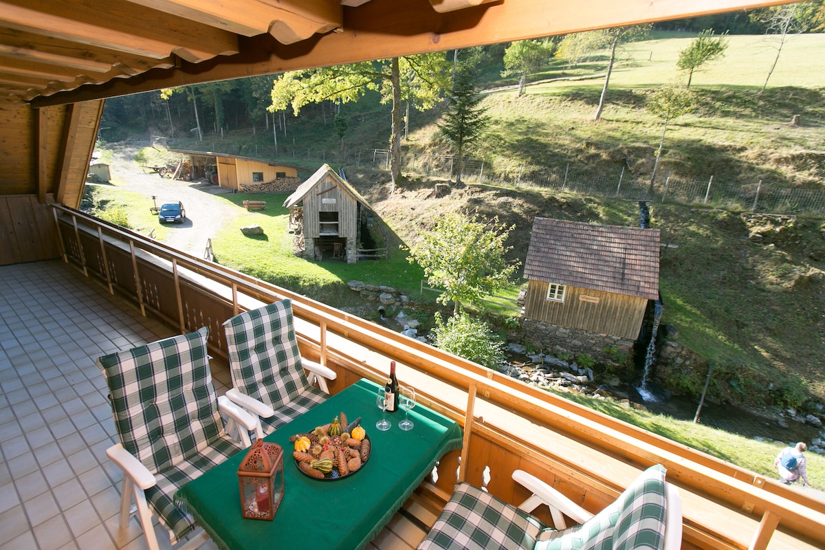 A spacious balcony is furnished with two checkered chairs and a green table, set with an assortment of local snacks. The view reveals a winding path and a gently flowing stream, bordered by rustic buildings and lush greenery in the distance.