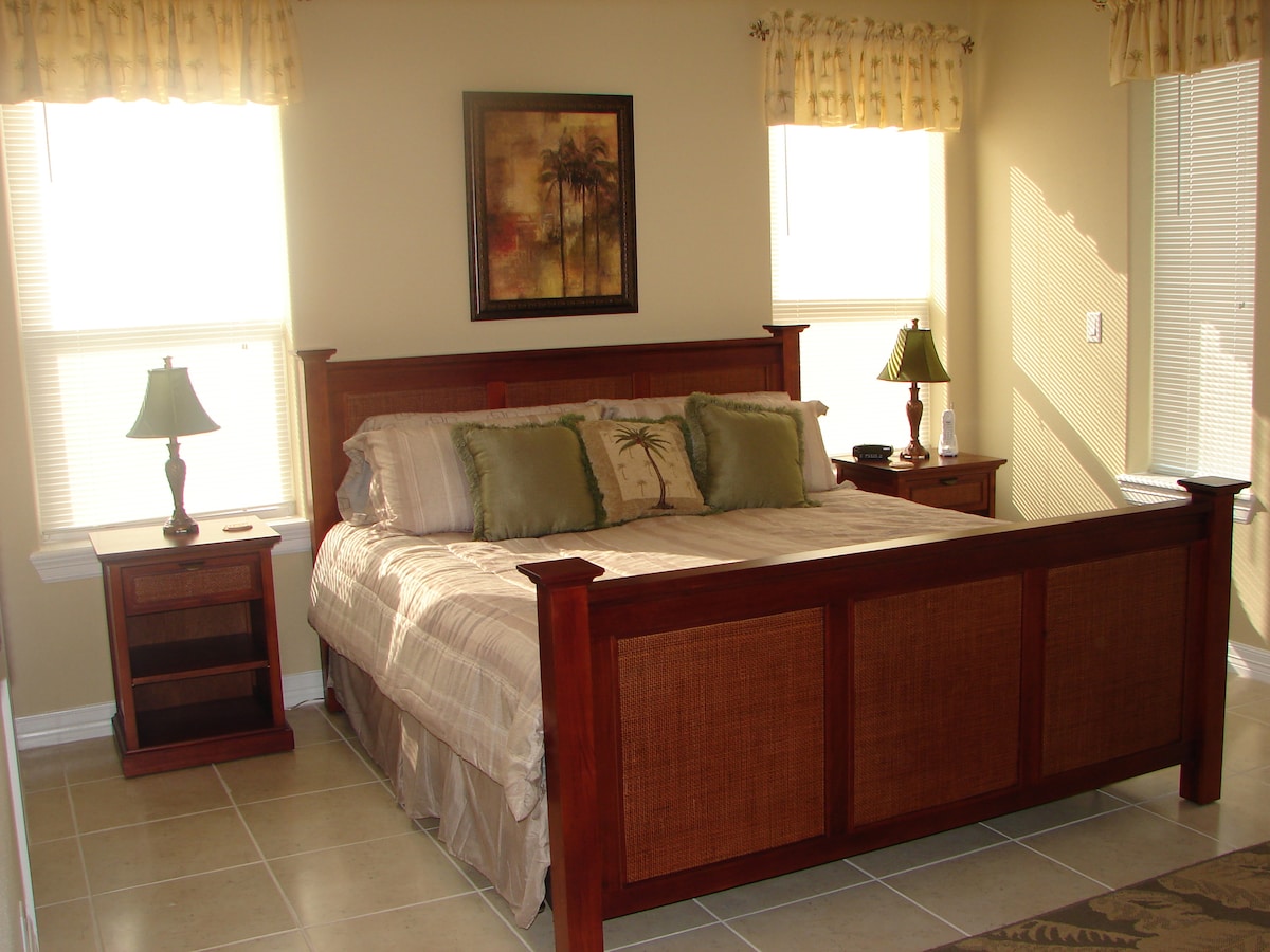 A spacious bedroom features a wooden bed frame with a neutral-toned bedding set and decorative pillows. Two bedside tables with lamps are positioned on either side, while natural light streams through large windows adorned with soft, light-colored curtains.