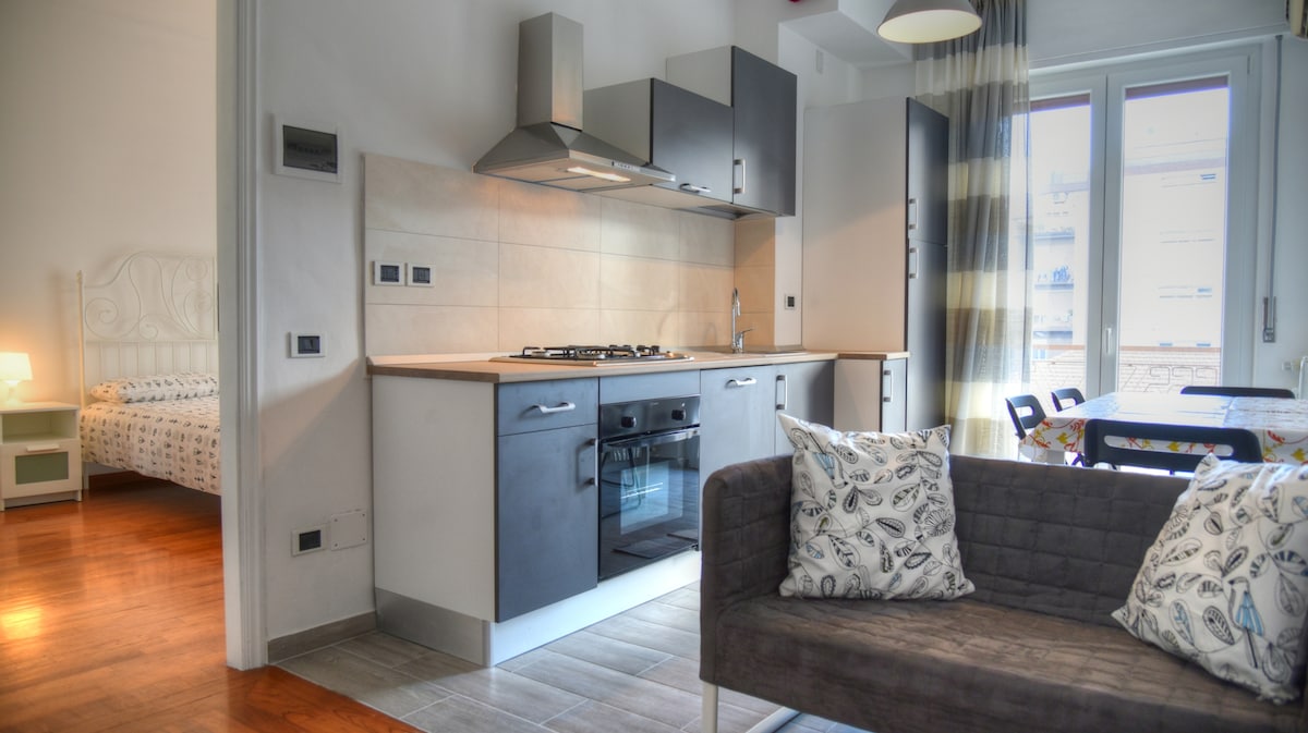 A modern kitchen area features a gas stovetop and sleek cabinetry, complemented by a cozy sitting area with patterned cushions. Nearby, a light-filled space includes a dining table visible through large windows, leading to a separate bedroom space with a bed framed against a soft-colored wall.