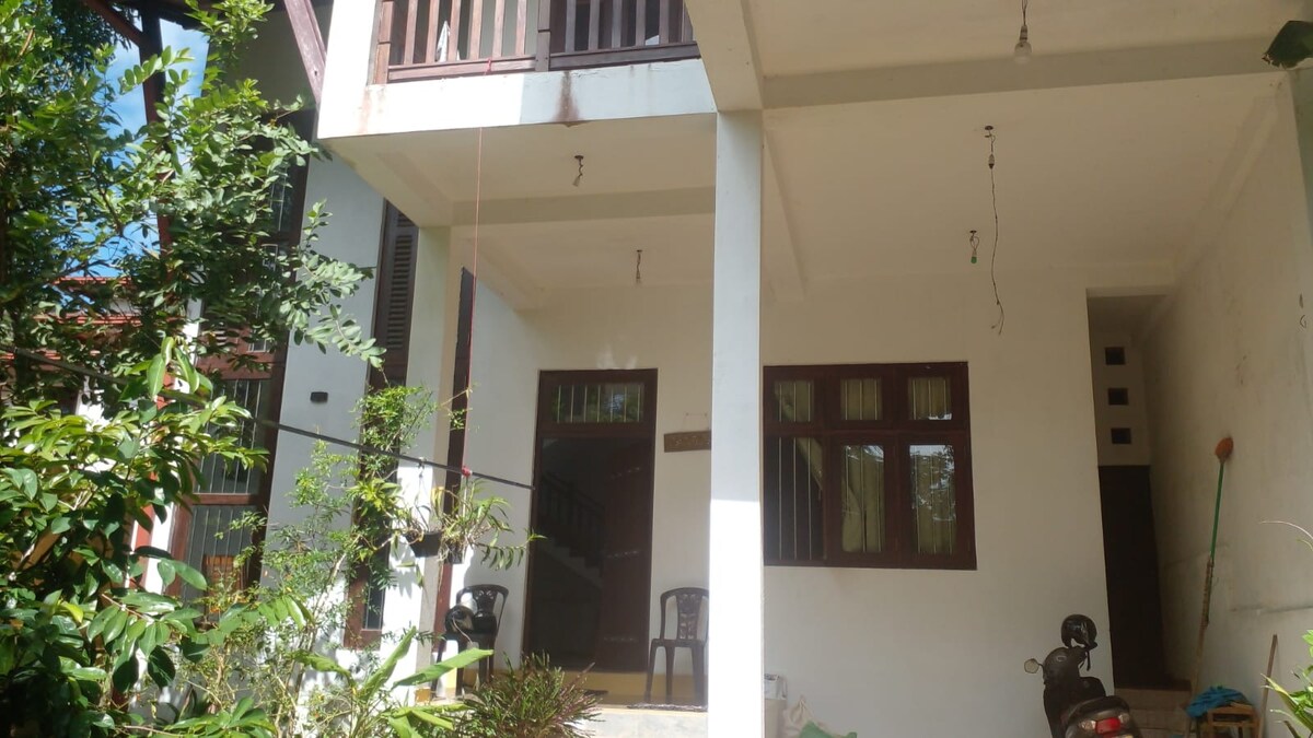 A two-story entrance area is framed by greenery, showcasing white walls and large windows that allow natural light to fill the space. A small seating area and a bike are visible, contributing to a welcoming atmosphere.
