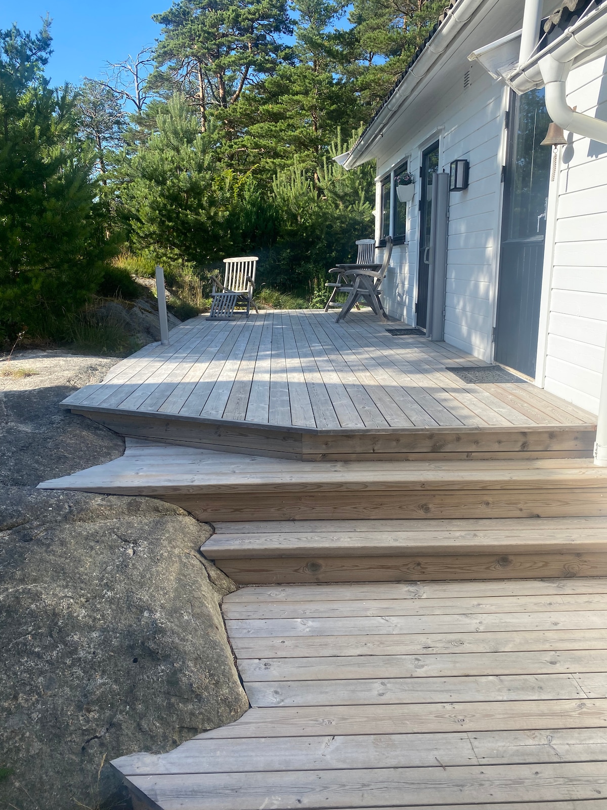 A wooden deck extends from the entrance of the property, featuring a smooth surface and a slight overhang. A rustic wooden chair and a small table are positioned on one side, surrounded by greenery and natural rock formations.