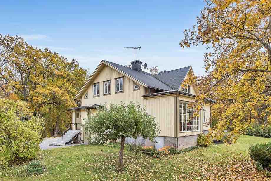 A charming two-story villa is set amidst vibrant autumn foliage, featuring large windows and a classic design. The surrounding landscape includes lush greenery, with a pathway leading to an entrance that is framed by trees, enhancing the peaceful country atmosphere.