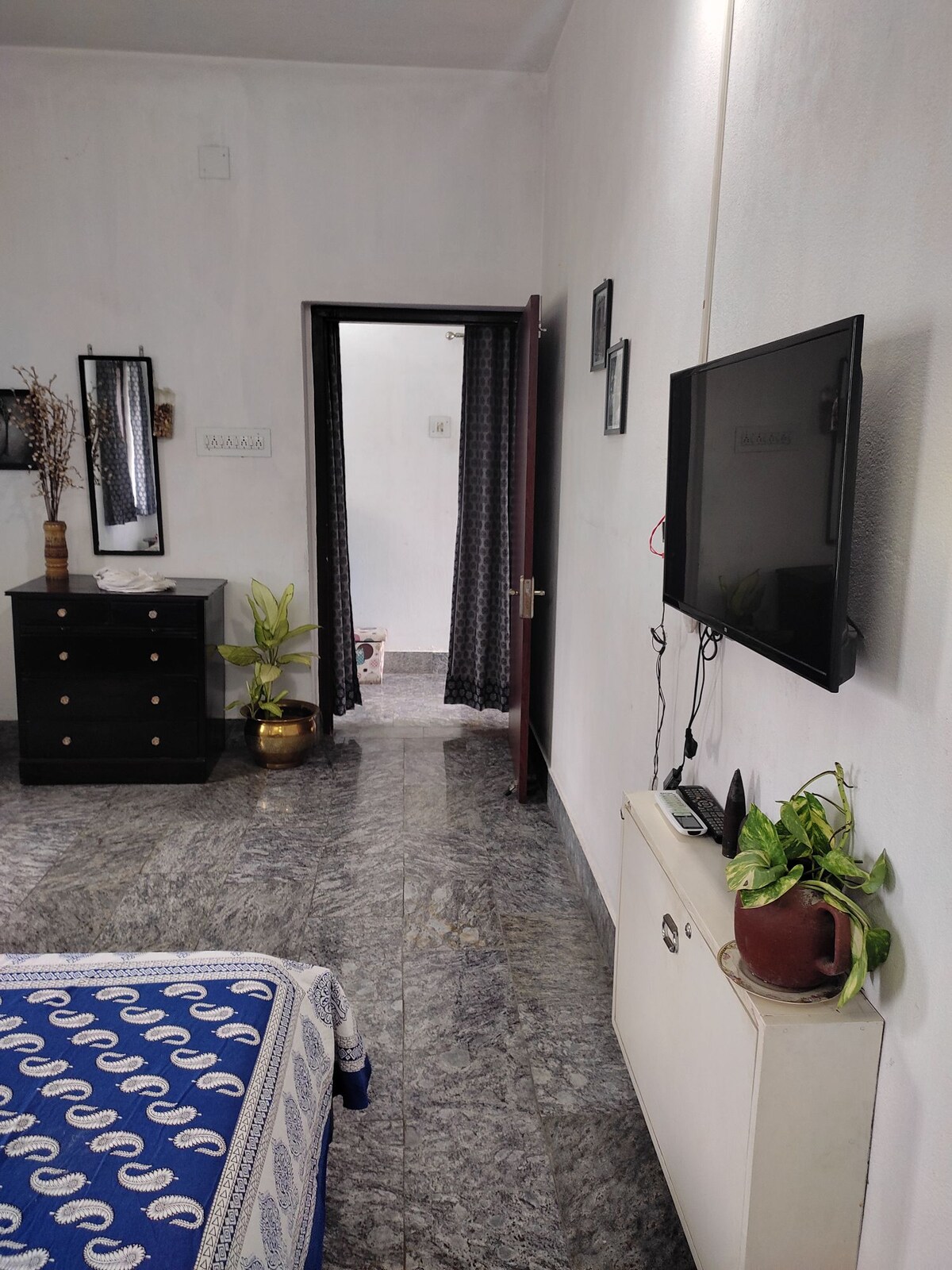 A well-lit room features a stylish dark dresser and a flat-screen television mounted on a neutral wall. A decorative plant sits on a white cabinet, while a doorway leads to an adjoining space, enhancing the room's privacy.