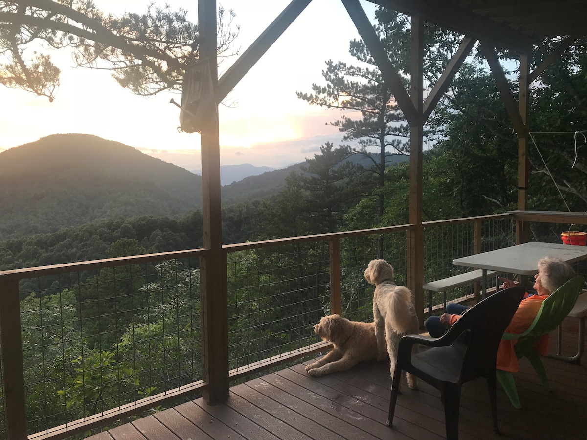 A spacious deck features a table, chairs, and a stunning view of the mountains and forest. Two dogs are visible, enjoying the serene sunset. The natural surroundings, including trees and rolling hills, create a peaceful and private atmosphere.