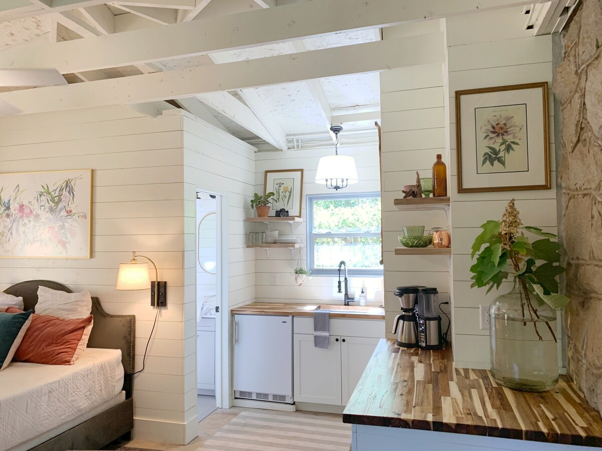 The interior features a cozy sleeping area with a spacious bed adorned with colorful cushions. A kitchenette is seen with wooden countertops, shelves, and a coffee maker. Natural light filters in through the window, illuminating the soft color palette and rustic design elements.