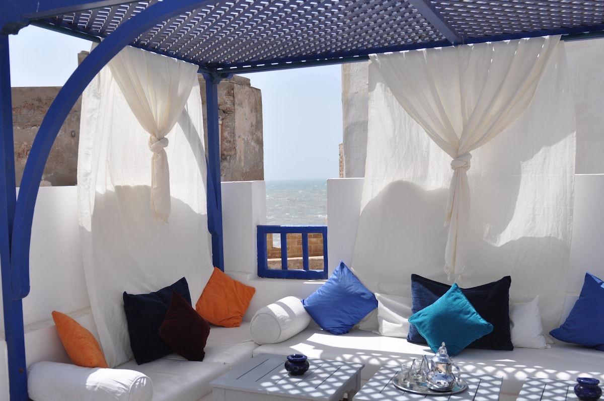 A shaded terrace features a white lounge area surrounded by sea views. Soft, colorful cushions in orange and navy blue are arranged on the seating, with light fabric draping elegantly from the structure above, creating a relaxing environment for enjoying the ocean breeze.