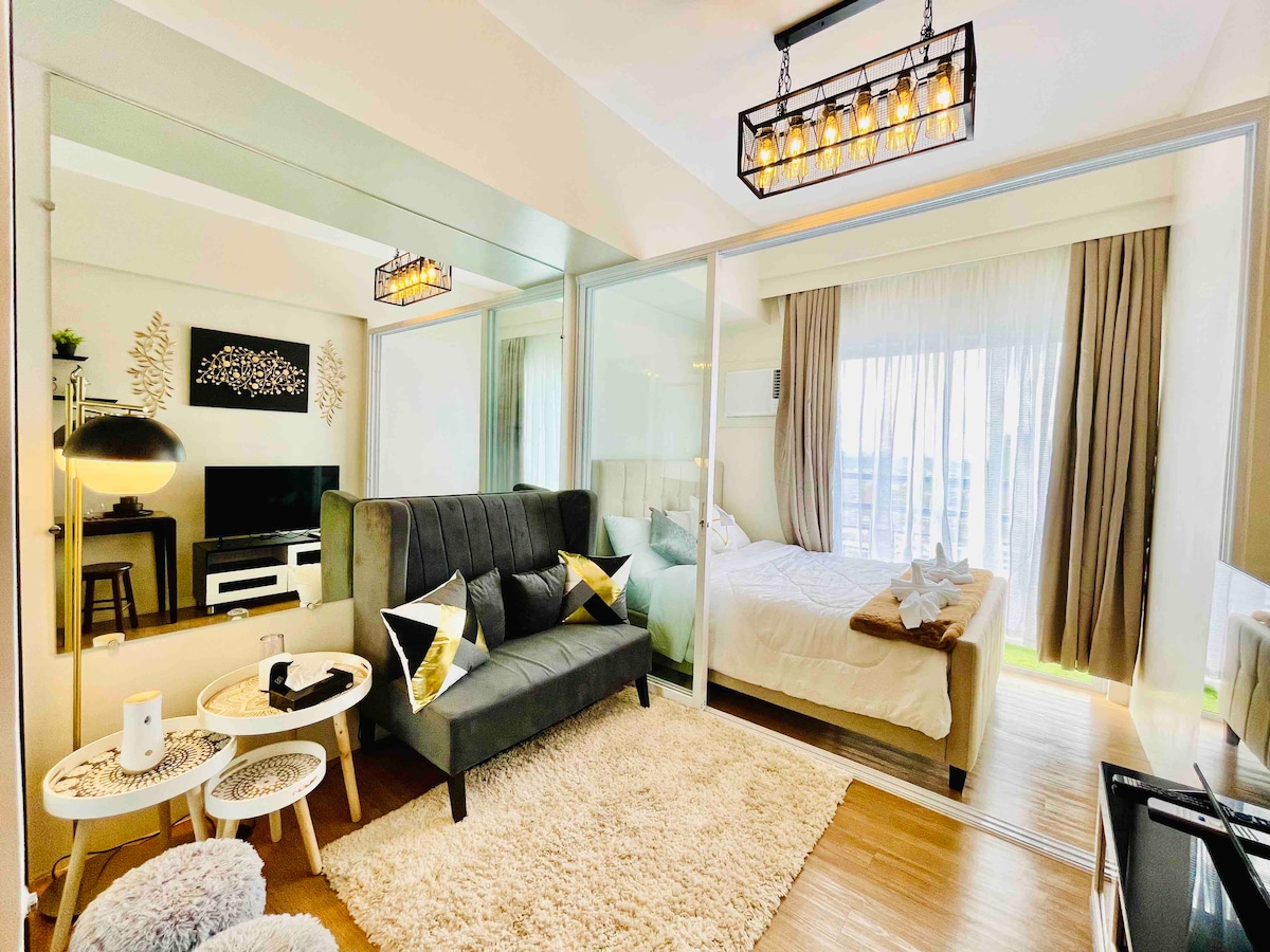 A modern studio unit is organized into a cozy living area and sleeping space. A gray sofa is complemented by decorative cushions, while a glass table sits nearby. The sleeping area features a spacious bed with soft linens. Natural light filters in through sheer curtains.