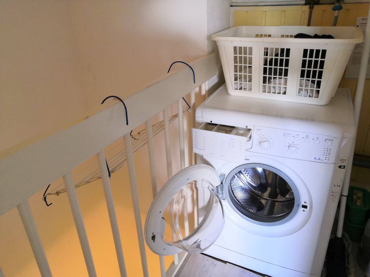 A washing machine is positioned next to a neatly arranged laundry basket filled with clothes. The machine's door is open, revealing the drum inside. Clothes drying spaces are available nearby, contributing to the laundry area’s functionality.