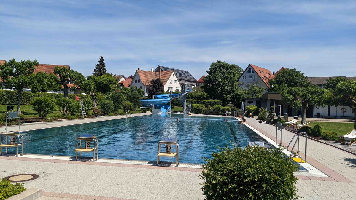 An outdoor swimming pool is shown, featuring a spacious layout with varying depths marked by numbers on the poolside. Surrounding greenery includes well-maintained bushes and trees. Lounge chairs are arranged along the edges, and nearby buildings with red-roofed tops provide a scenic backdrop.