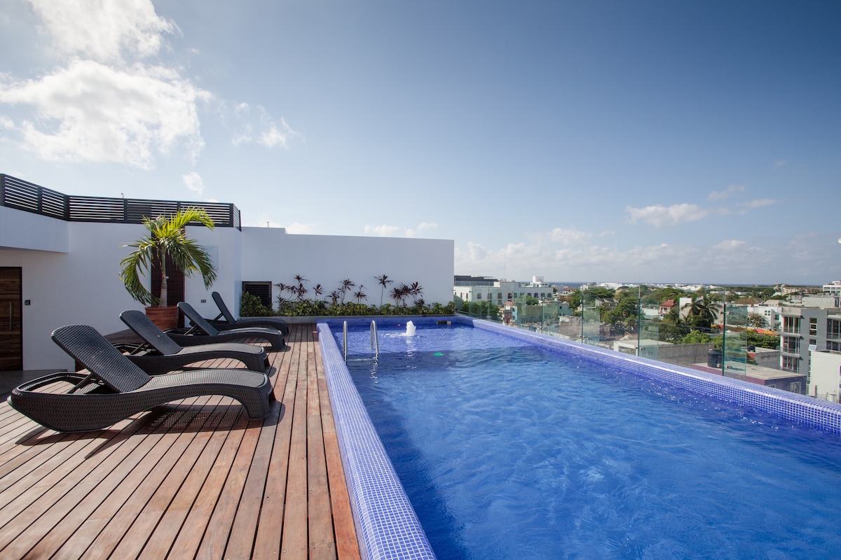 A rooftop swimming pool is surrounded by wooden decking and lounge chairs, offering a view of the skyline. Clear blue waters reflect the sky above, with lush greenery positioned near the pool's edge, creating a serene retreat.