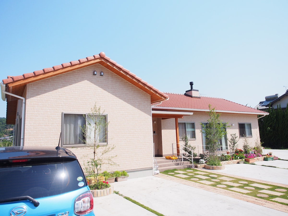 A single-story house is shown, featuring a light brick exterior and a red-tiled roof. The front lawn is landscaped with neat greenery and flower beds. A driveway is visible, with a parked blue vehicle positioned near the house, providing a welcoming access point.