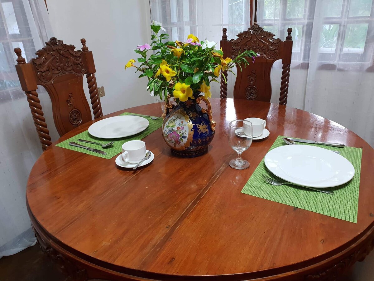 A round wooden dining table is set with two white plates and cups, complemented by green placemats. A vibrant floral arrangement in a decorative vase adds color to the center, while intricately designed wooden chairs surround the table, creating an inviting dining space.