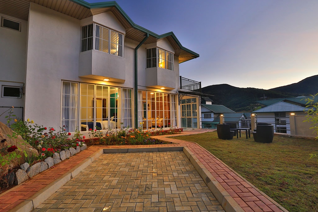 The exterior of the bungalow is presented, featuring a well-maintained garden bordered by colorful flowers. Large glass windows reflect the evening light, and a paved pathway leads to a seating area with modern chairs. Rolling hills create a serene backdrop.
