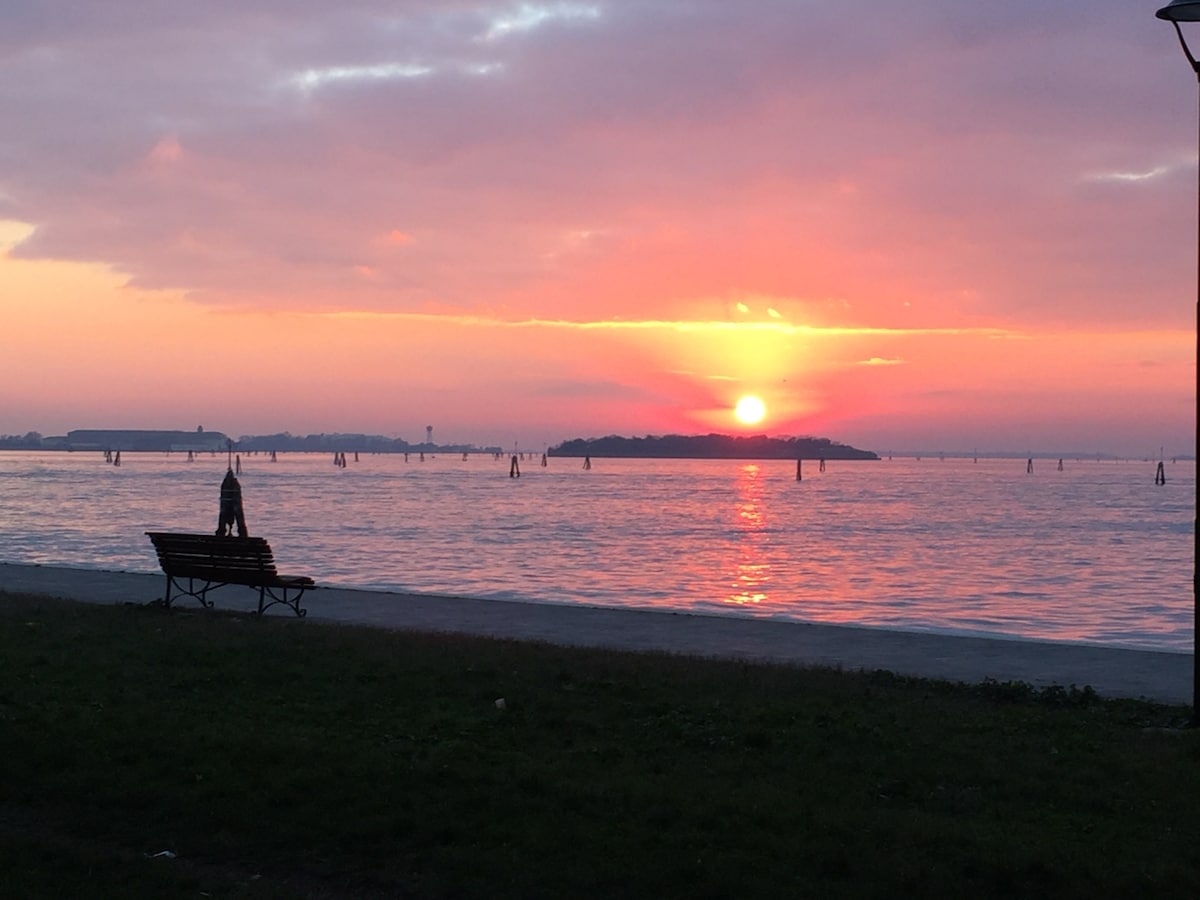 A serene view of the water stretching towards the horizon, where the sun is setting in shades of orange and pink. A lone bench is positioned in the foreground, offering a peaceful spot to enjoy the surroundings, while gentle waves reflect the colorful sky.