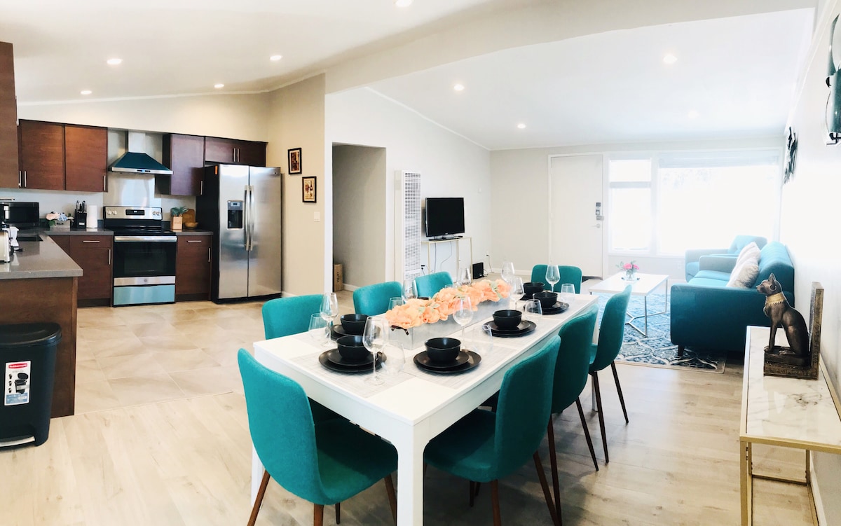 An open living space features a dining area with a white table set for six, surrounded by teal chairs. The kitchen showcases modern appliances, including a stainless steel refrigerator and gas stove. A comfortable seating area is visible, with a light-colored sofa and a television in the background.