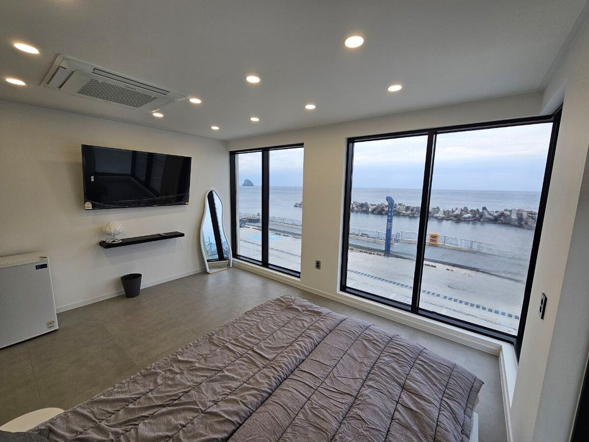 A bright room features floor-to-ceiling windows offering an unobstructed view of the ocean. A spacious bed is complemented by modern decor, including a large flat-screen TV and a full-length mirror. Natural light fills the space, creating an open and airy atmosphere.