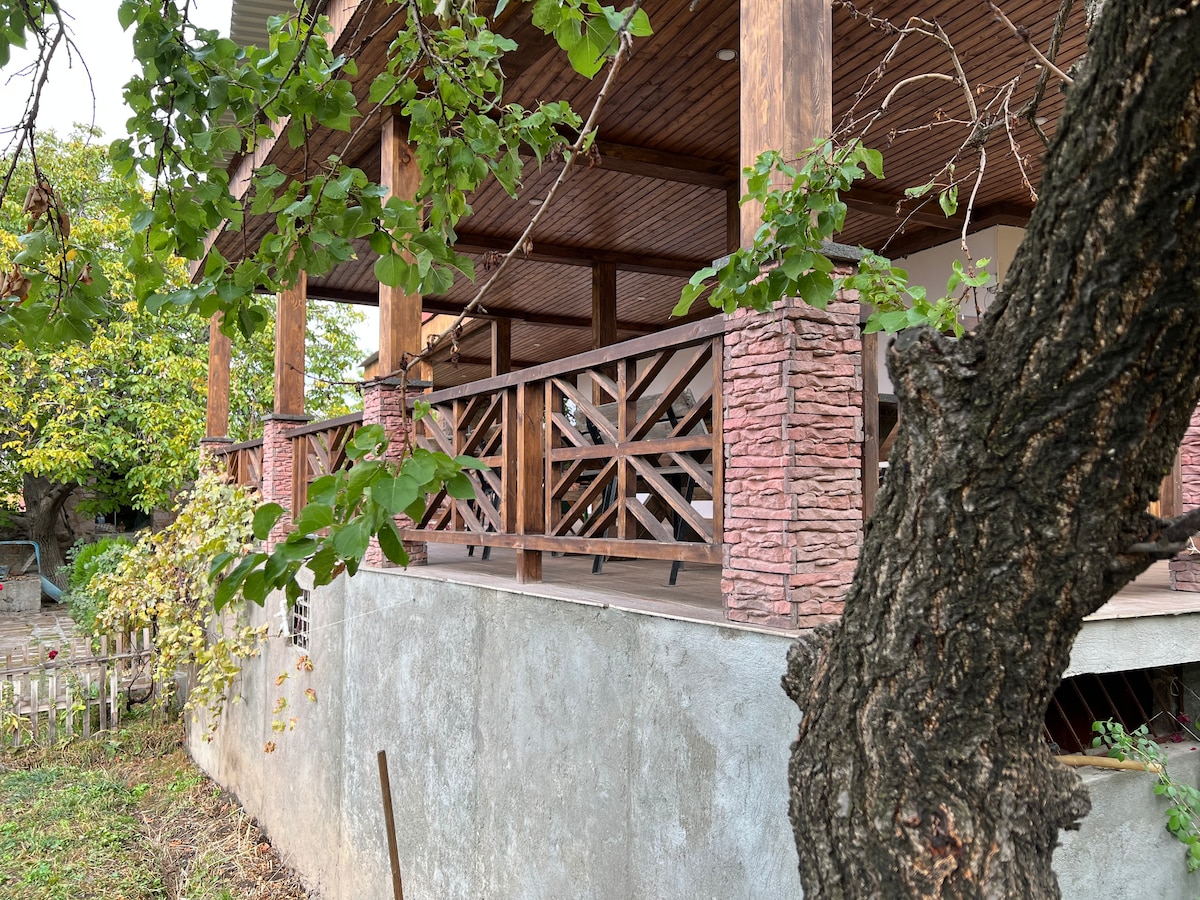 A spacious porch area is supported by wooden pillars and adorned with a decorative stone railing. The wood ceiling enhances the structure's natural aesthetic, while lush greenery surrounds the space, creating a tranquil outdoor retreat.