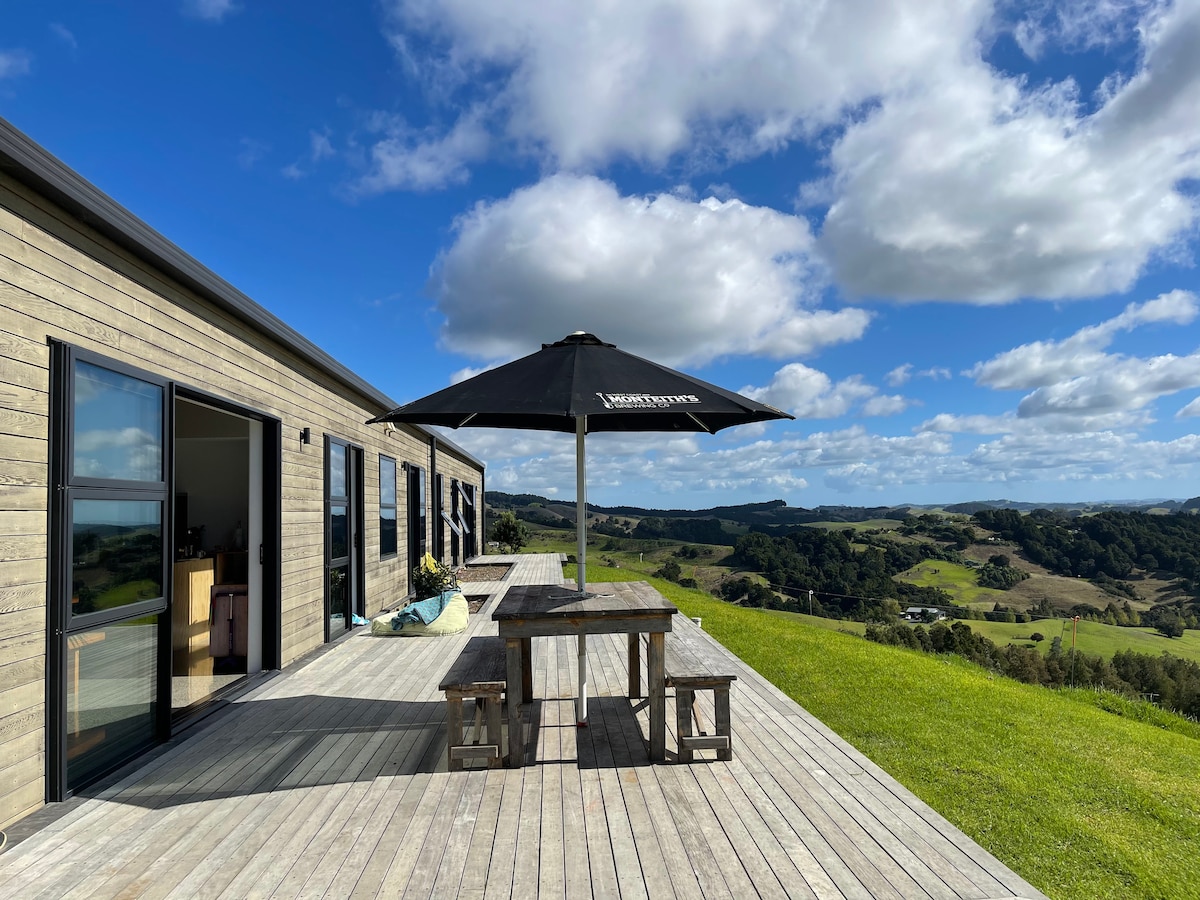 An expansive deck overlooks a scenic rural landscape, featuring a wooden table and benches shaded by a large umbrella. Large glass doors provide access to the interior, enhancing the connection between indoor and outdoor spaces, all set against a backdrop of rolling hills and a blue sky.