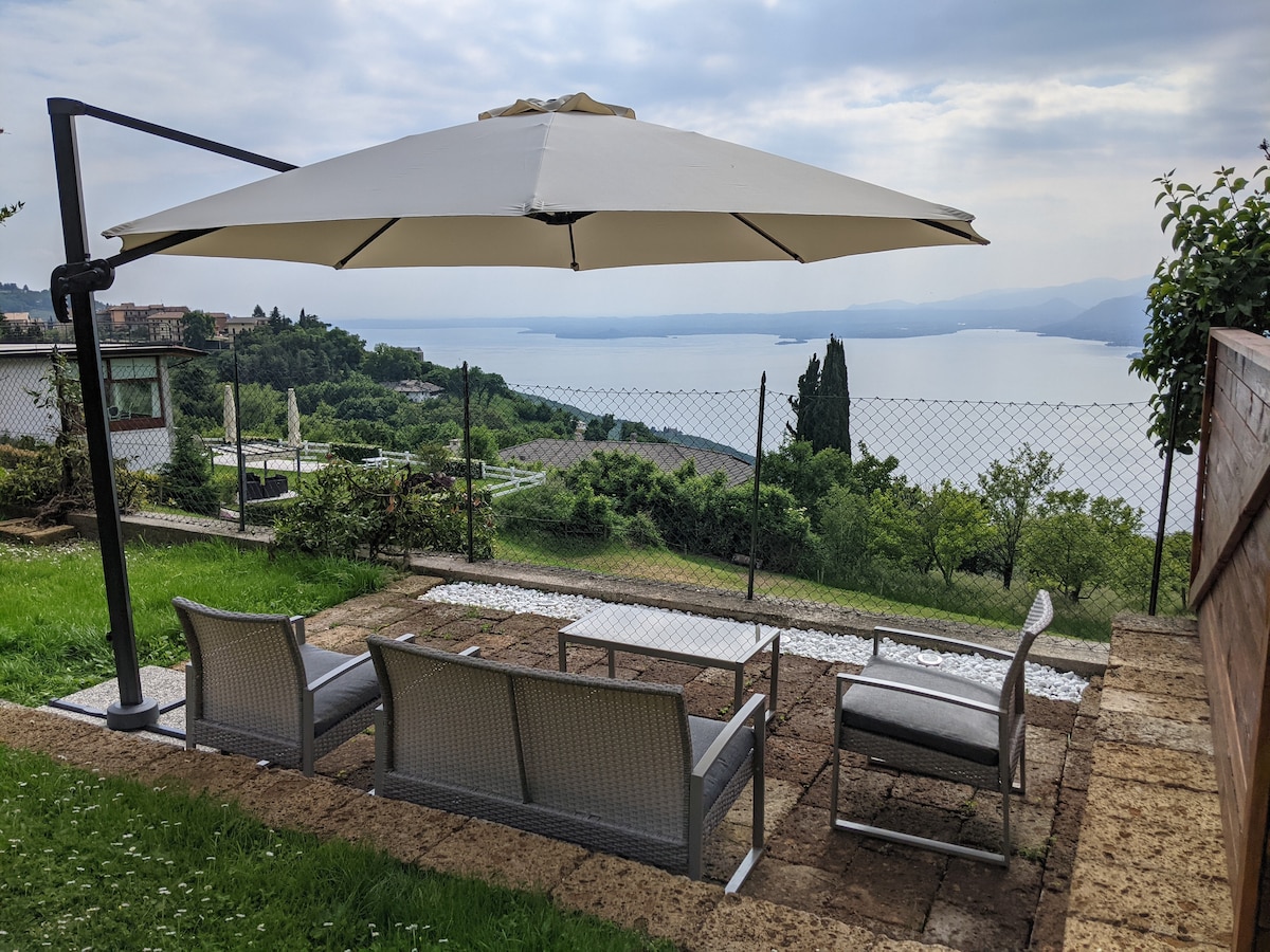 An outdoor seating area is highlighted under a large umbrella, overlooking a panoramic view of Lake Garda. The space features comfortable lounge chairs and a small table, set on a stone patio surrounded by greenery, providing a serene environment for relaxation.