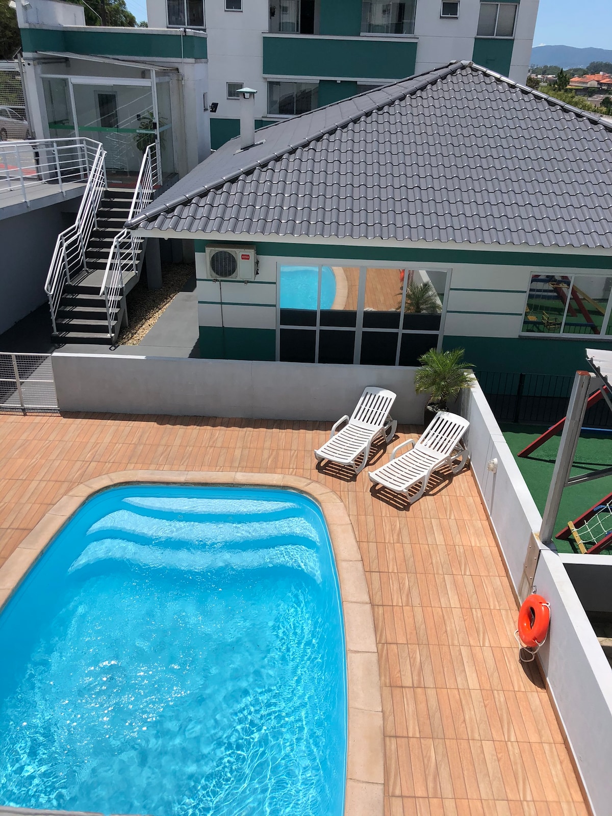 An inviting outdoor space features a swimming pool surrounded by a wooden deck. Two lounge chairs are positioned nearby, with the exterior of a nearby building in view, showcasing a modern architectural style and ample natural light.