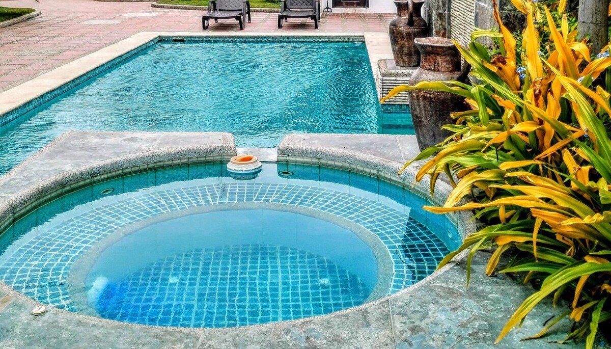 A tranquil pool area features a clear blue swimming pool and a circular jacuzzi surrounded by natural stone. Lush yellow and green foliage adds vibrancy to the setting, while terracotta pottery decorates the space, enhancing the peaceful ambiance.