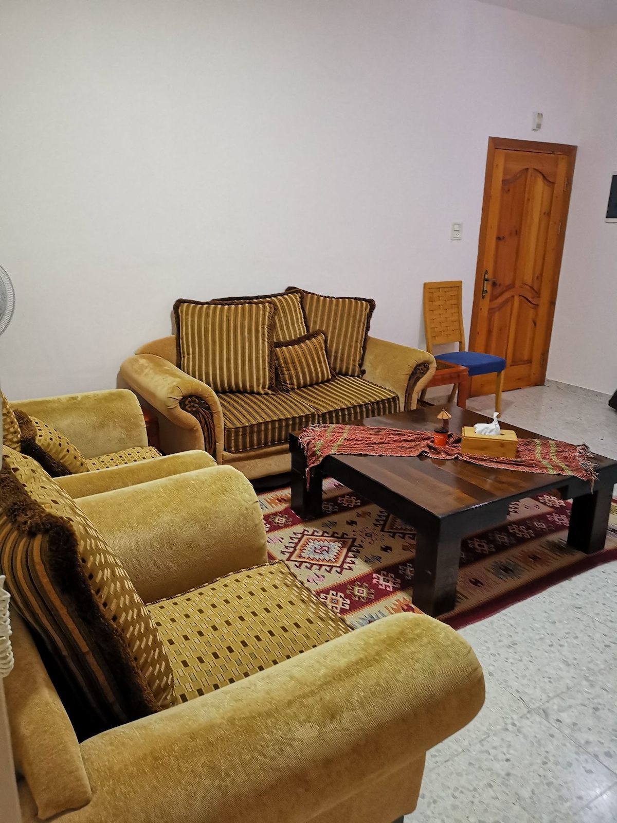 A seating area features a plush beige sofa with decorative cushions, complemented by two matching armchairs. A dark wooden coffee table occupies the center, adorned with a decorative tray. A colorful area rug lies underneath, and a wooden door is visible at the rear.