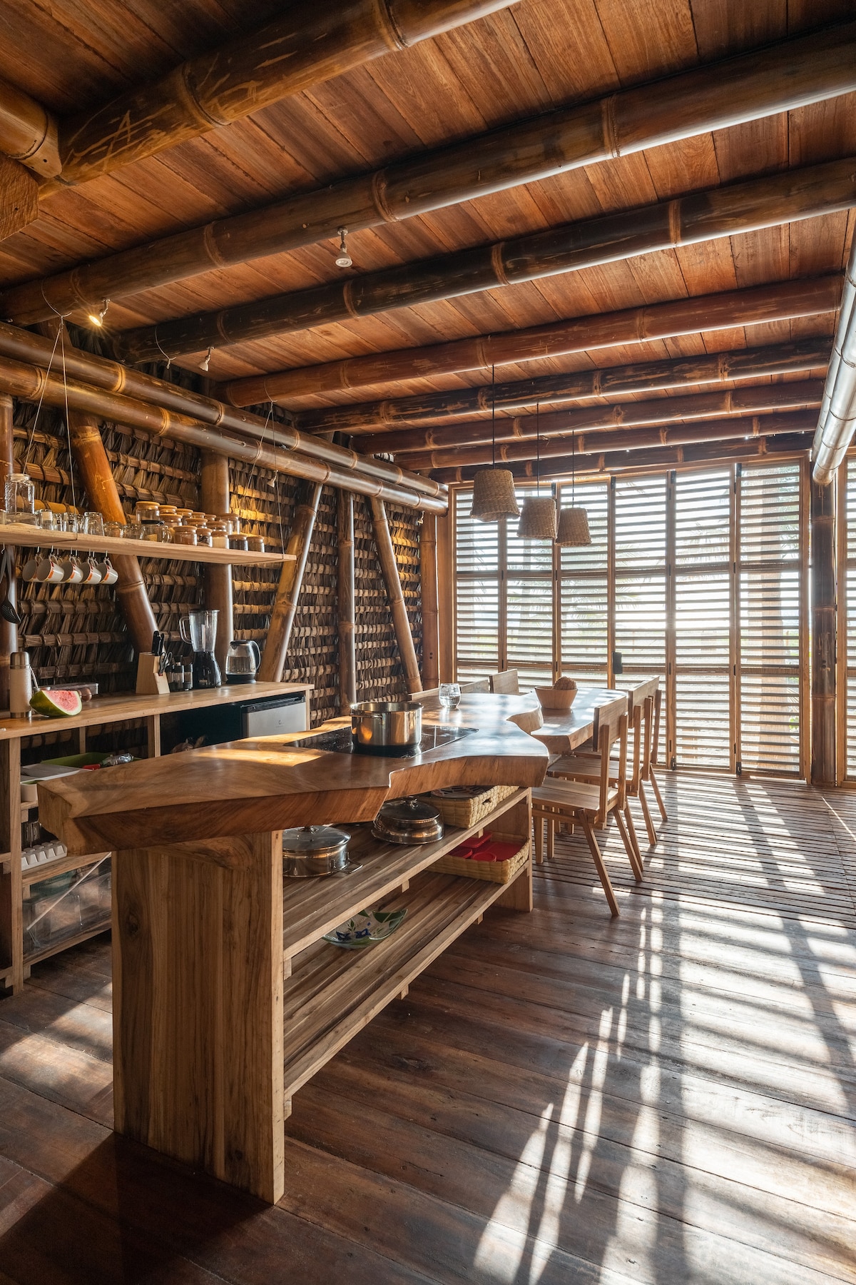 A spacious kitchen area features wooden cabinetry and a long dining table surrounded by chairs. Natural light filters through slatted windows, creating a pattern of shadows on the wooden floor. Shelves display glassware and kitchen essentials, highlighting the open design.