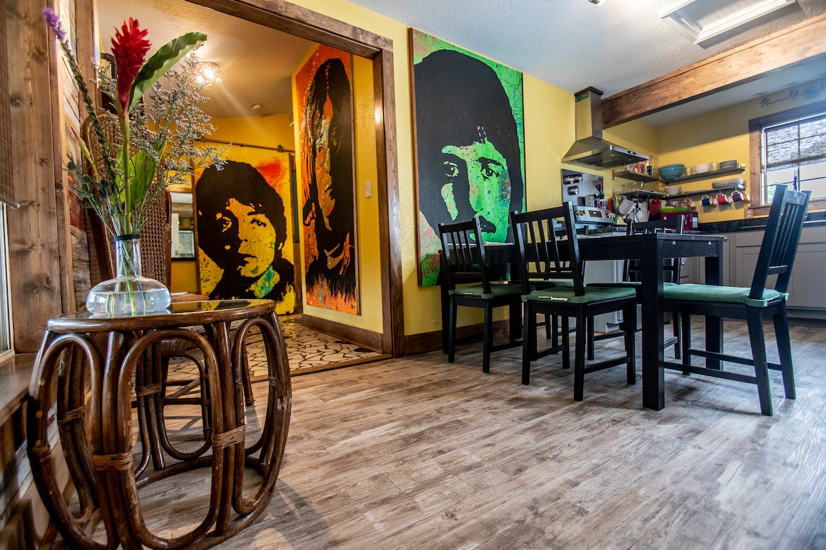 A vibrant dining area features a round table surrounded by four black chairs with green cushions. Colorful artwork, including large portraits on yellow walls, adds a lively atmosphere. A well-equipped kitchen is visible in the background, with modern appliances and open shelving.