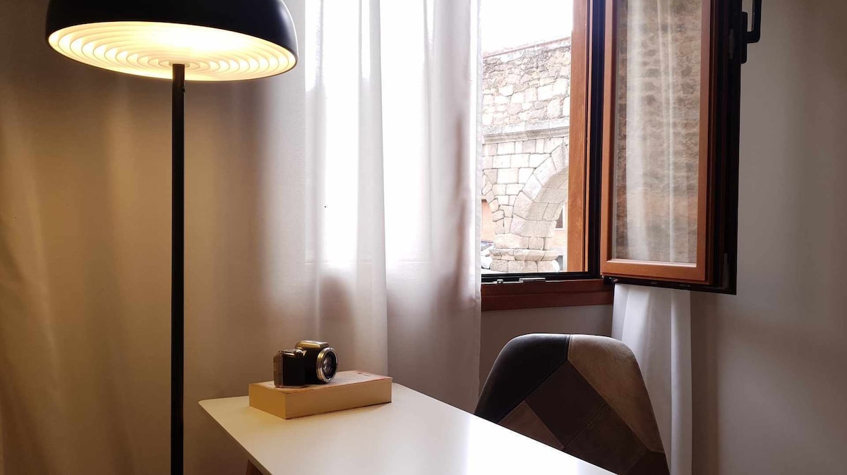 A well-lit workspace features a white desk and a comfortable chair, positioned near a window adorned with sheer curtains. Natural light enters the room, highlighting a vintage camera placed on the desk and offering a view of the outside architecture.