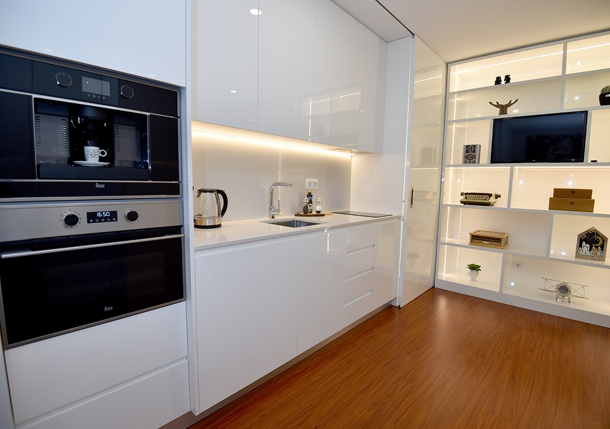 A modern kitchen is showcased, featuring sleek white cabinetry and integrated appliances. A coffee machine and kettle are positioned on the countertop, complemented by a stainless steel sink. Illuminated shelving highlights decorative items, enhancing the room's contemporary design.